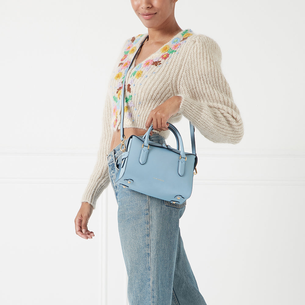 A woman in light blue jeans and a cream floral cardigan holds a pale blue structured handbag against a white background.
