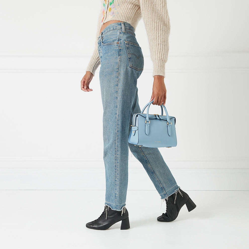 A woman in light wash jeans and black ankle boots carries a pale blue structured handbag against a white background.
