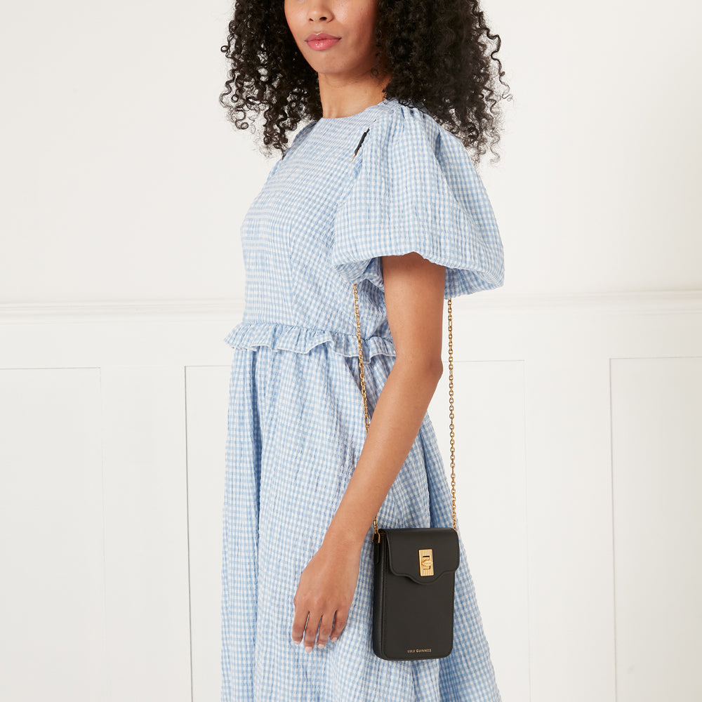 Woman in a light blue gingham dress holding a black Lily Douglas crossbody bag with a gold chain strap against a white background.
