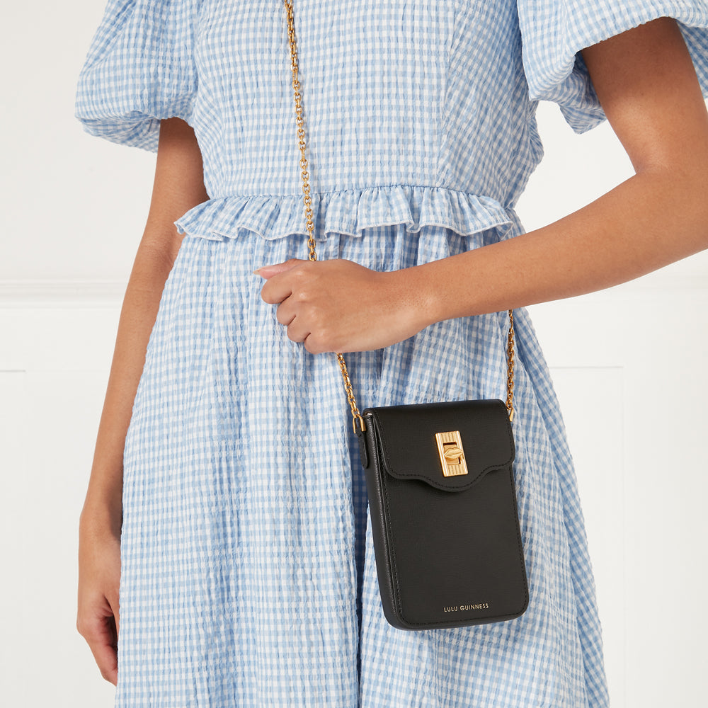 Black Lulu Guinness crossbody bag with gold chain strap and clasp, worn by a model in a light blue gingham dress against a white background.
