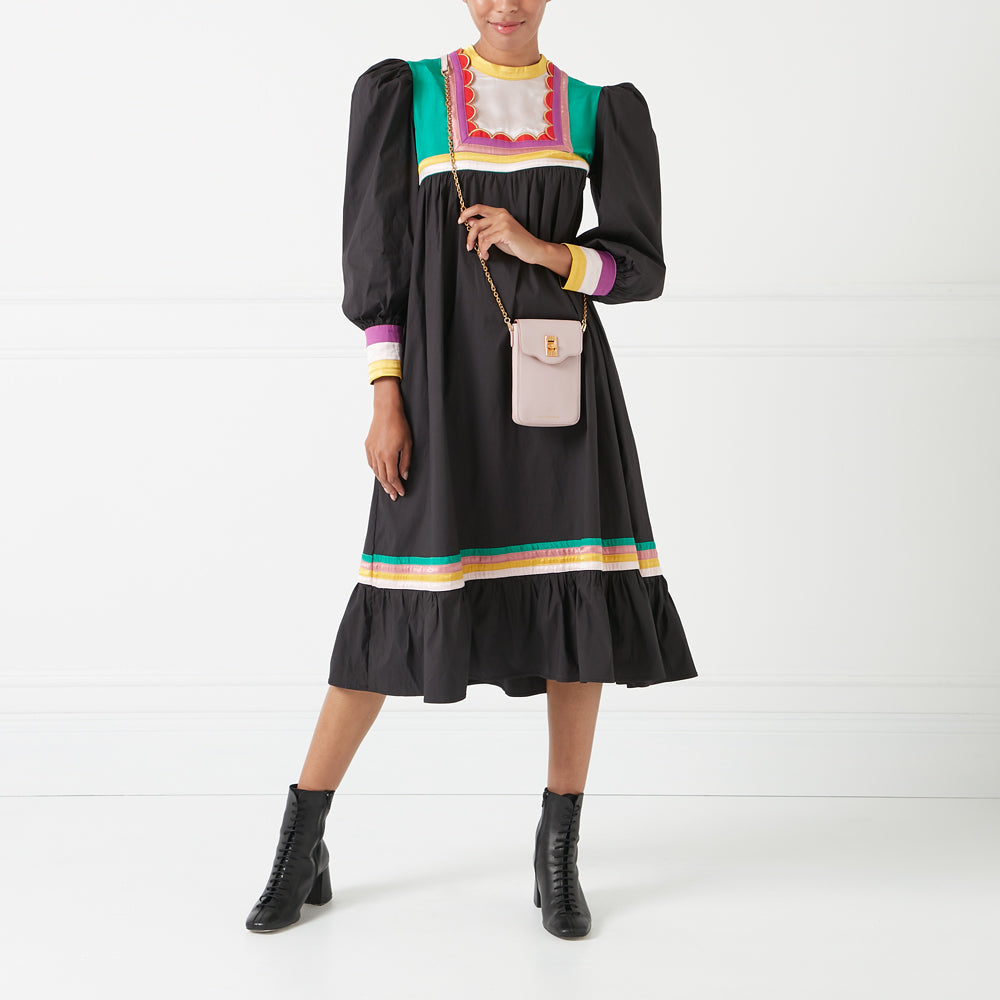 A woman in a black midi dress with colorful details and black ankle boots carries a light pink crossbody bag against a white background.
