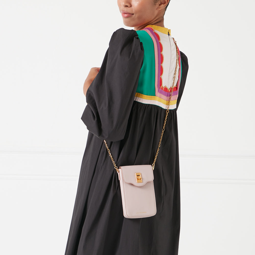A pale pink crossbody bag with gold hardware hangs from a gold chain on a model wearing a black dress with colorful details on the back.
