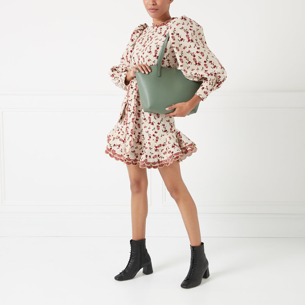 Woman in floral dress holding sage green tote bag against white background.
