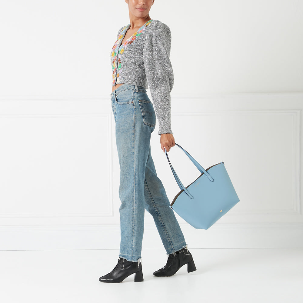 A woman in light wash jeans and a grey cardigan carries a light blue tote bag against a white background.
