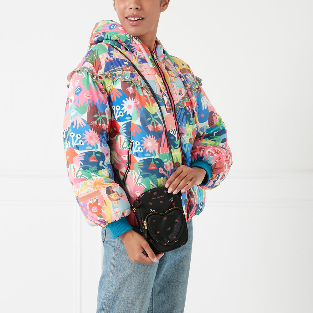 A model wears a colorful, floral puffer jacket and a black crossbody bag with a heart-shaped front pocket. The background is white.
