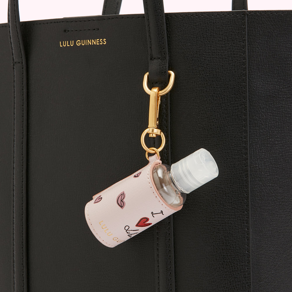 A black Lulu Guinness tote bag with a pink hand sanitizer holder attached to it. The hand sanitizer holder has a design of lips and hearts and says "I love Lulu Guinness" on it.