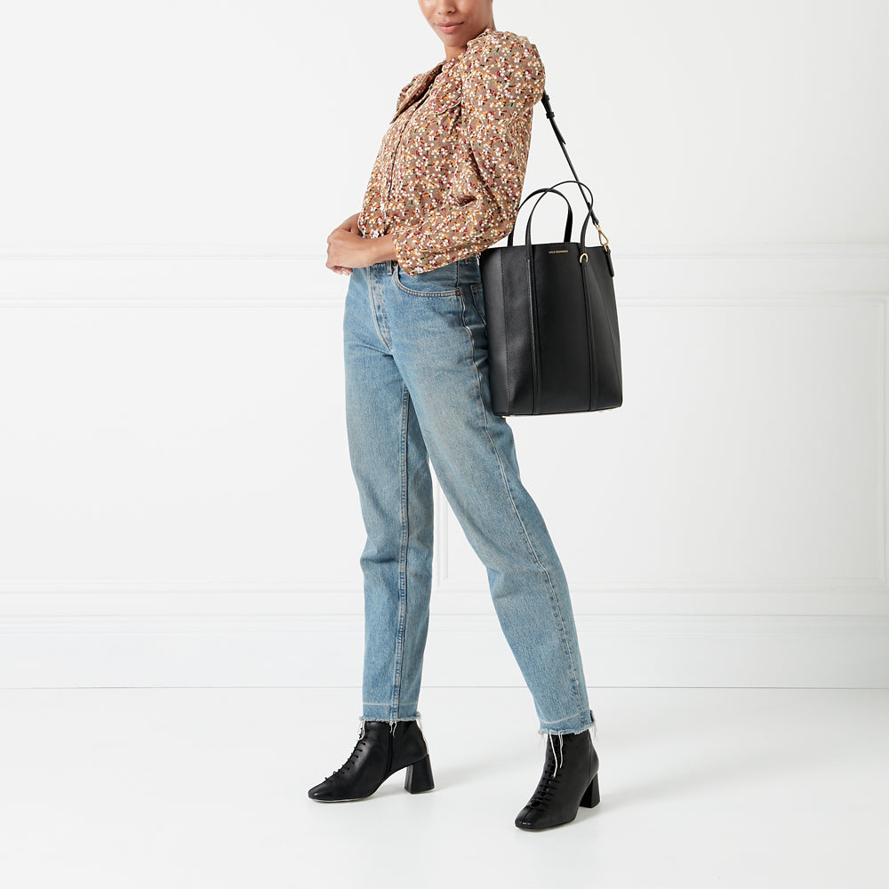 Model wearing light wash jeans, floral blouse, black ankle boots, and a large black tote bag against a white background.
