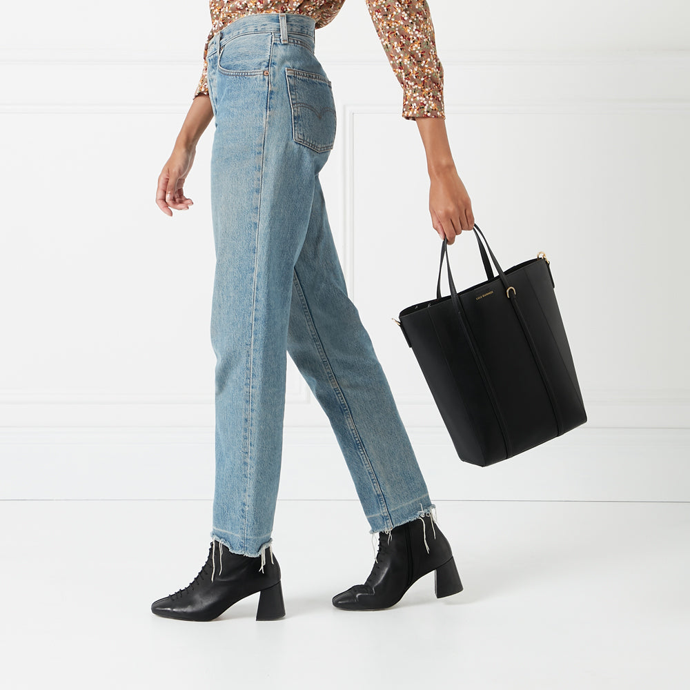 A woman in light wash jeans and black ankle boots carries a black tote bag against a white background.
