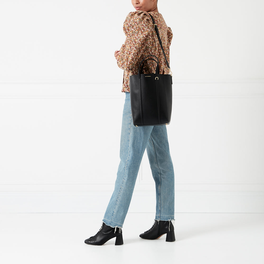 A woman in light wash jeans and a patterned top carries a black tote bag with a gold accent.