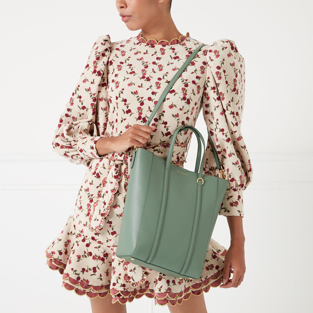 A woman in a floral dress holds a sage green tote bag.
