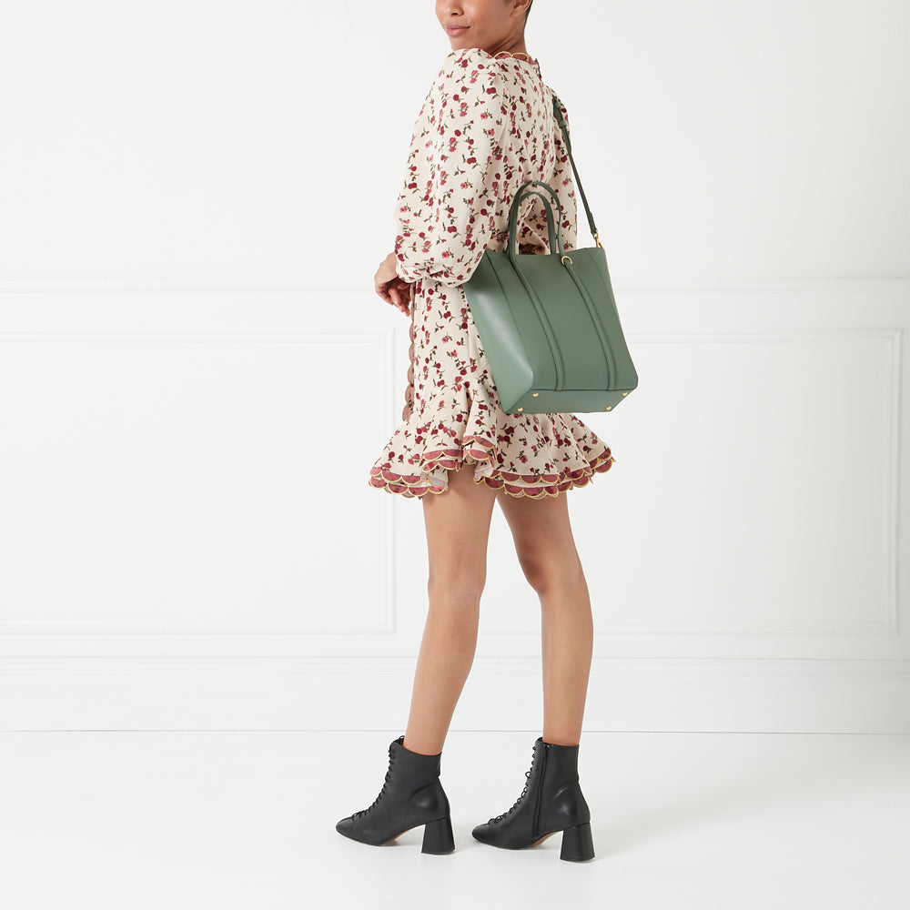 Woman in floral dress carrying a sage green tote bag and wearing black ankle boots against a white background.

