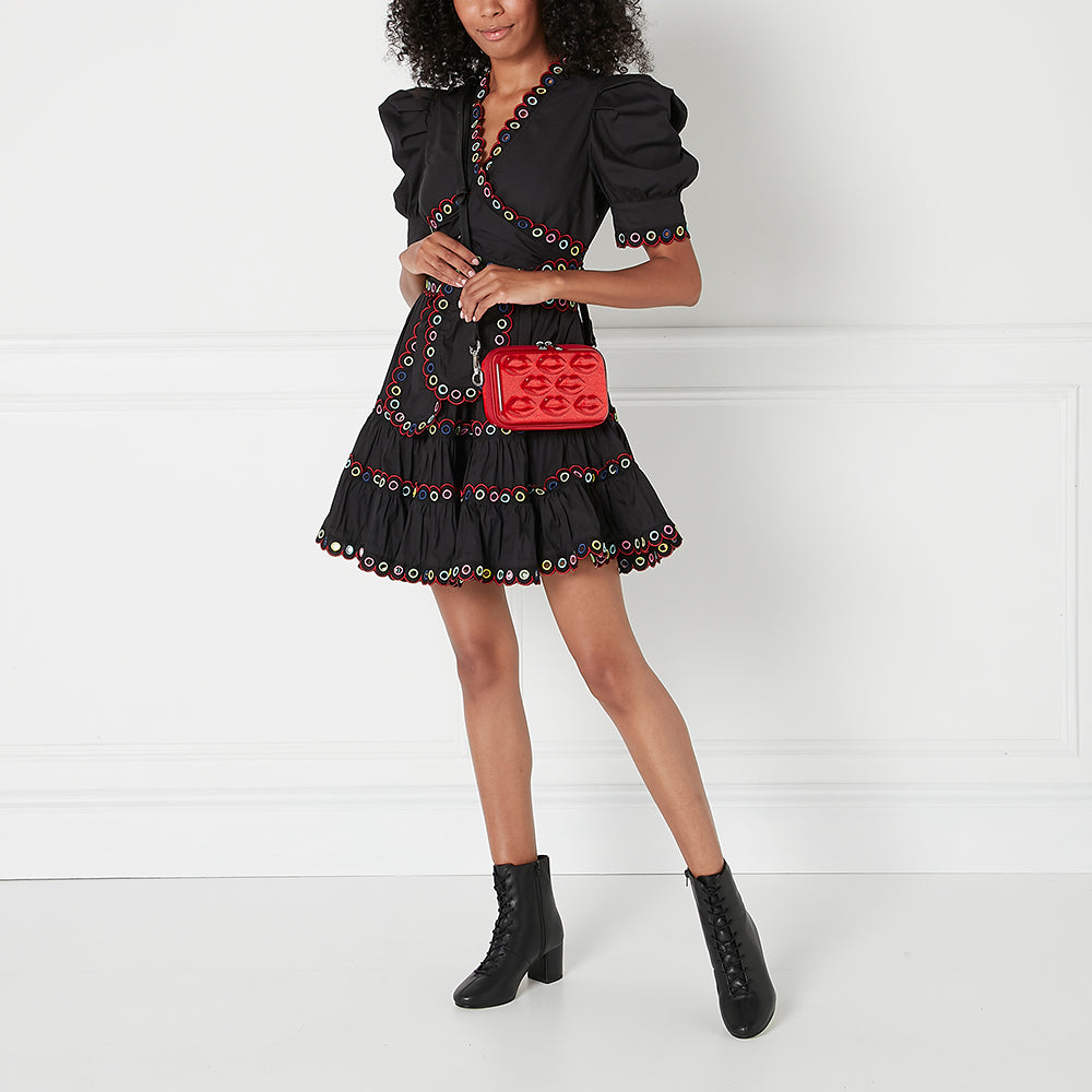A woman with curly hair models a black eyelet dress and black ankle boots while carrying a red lip-shaped crossbody bag against a white background.