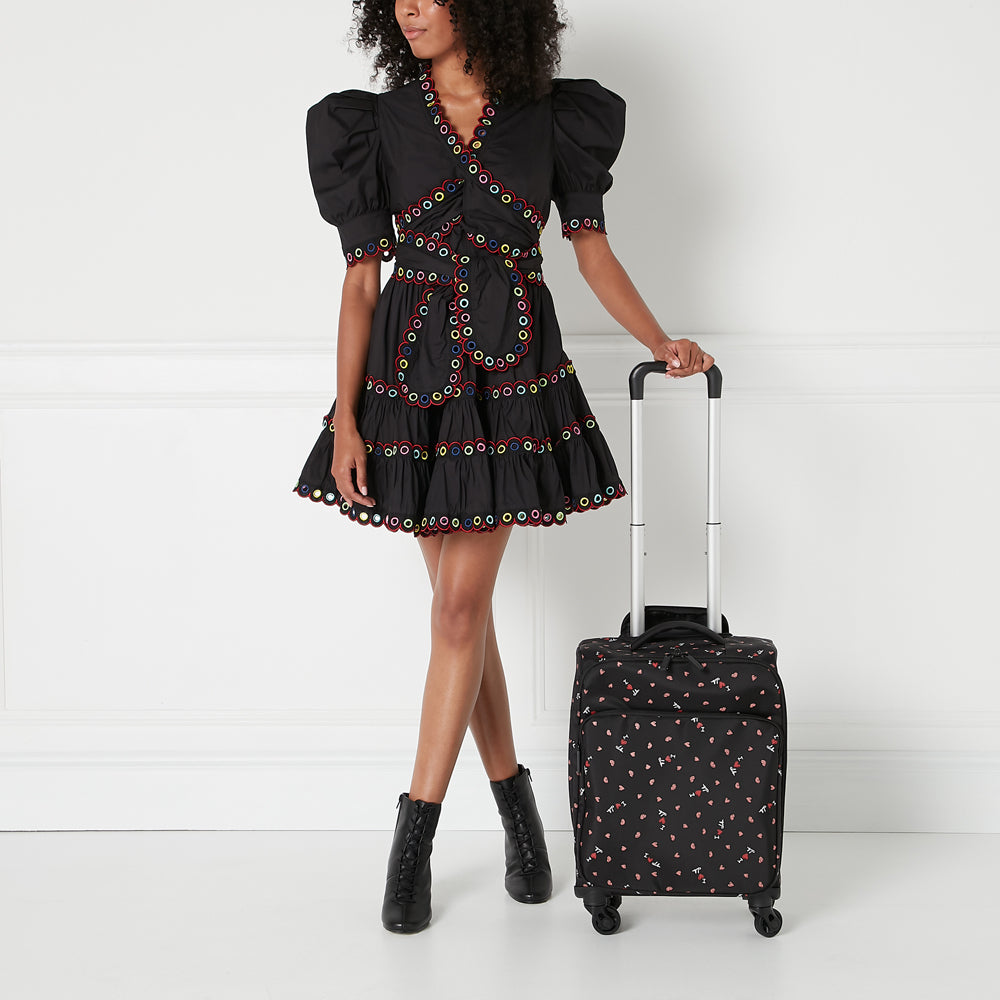 Woman in black eyelet dress and black ankle boots stands next to a small black rolling suitcase with a black and red heart print. The background is white.
