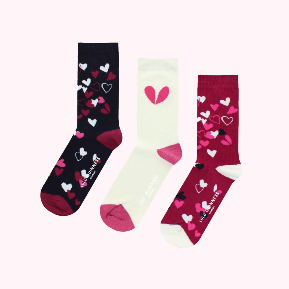 Three pairs of Lulu Guinness socks with heart designs; one pair is navy blue with pink hearts, one is off-white with a single broken pink heart, and one is burgundy with pink and white hearts.