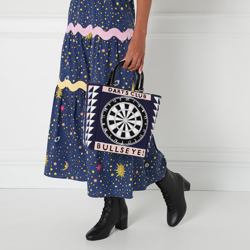 A woman in a navy blue midi dress with stars and moons holds a navy blue tote bag with a dartboard design. The bag has the words "Darts Club" and "Bullseye!" on it. The background is off-white.

