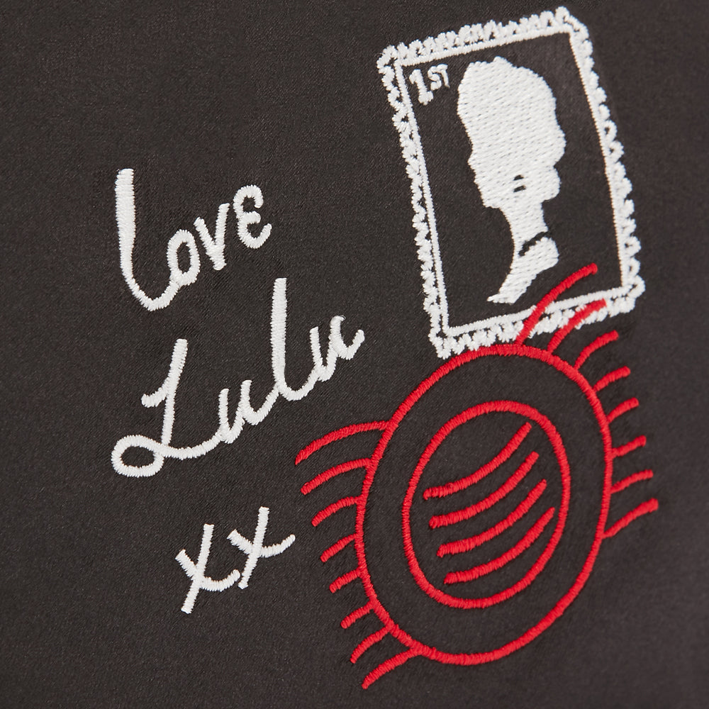 Close-up of black fabric with embroidered white cursive text "Love Lulu xx", a red sun-like design, and a white embroidered postage stamp.
