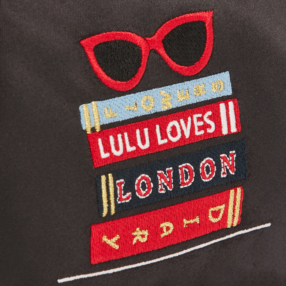 Close-up of black satin fabric with embroidered red sunglasses and a stack of books with text "Lulu Loves London" in different colors.