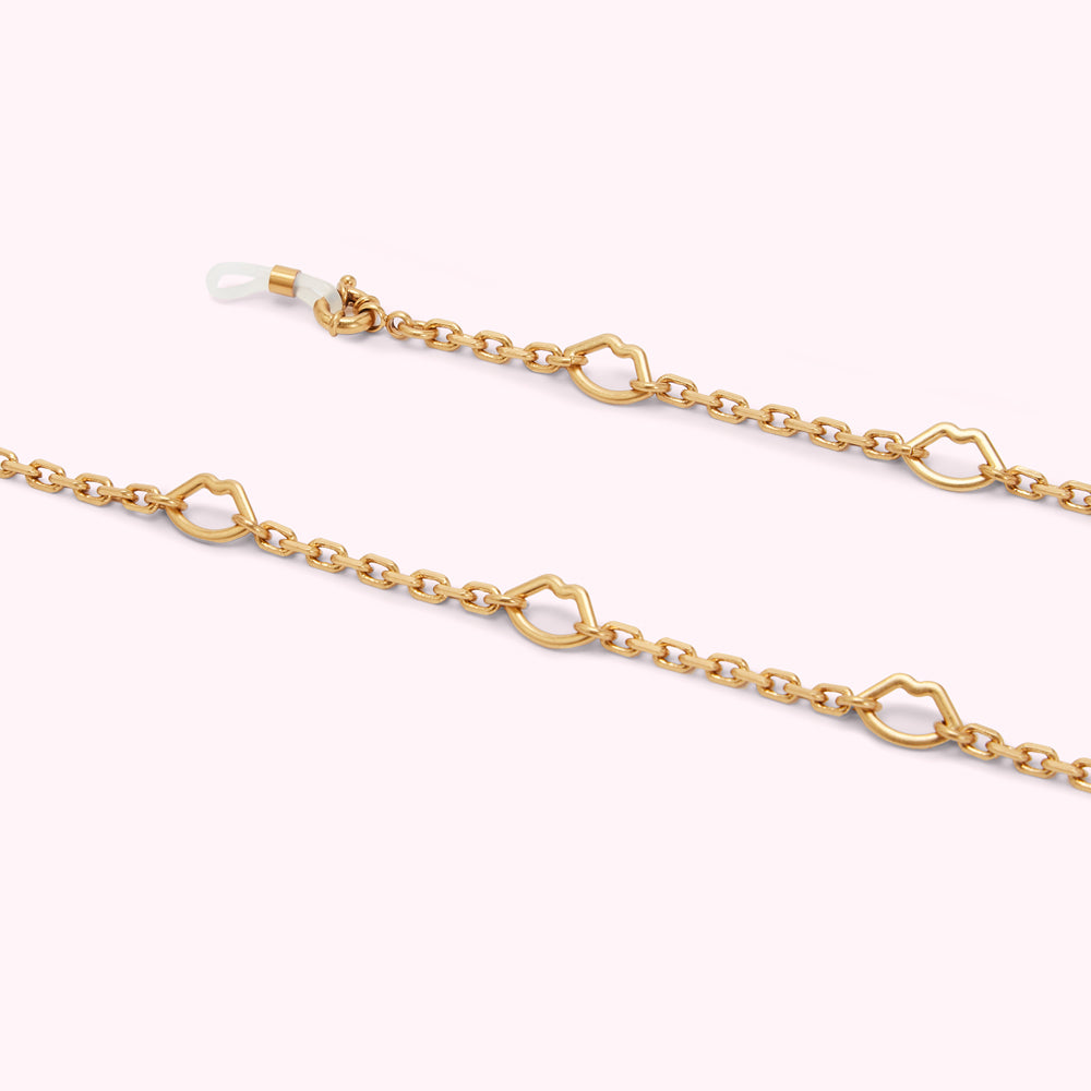 Gold eyeglasses chain with lip-shaped charms against a light purple background.
