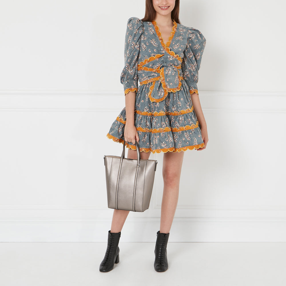 Woman in a floral dress and black boots holding a silver tote bag against a white background.
