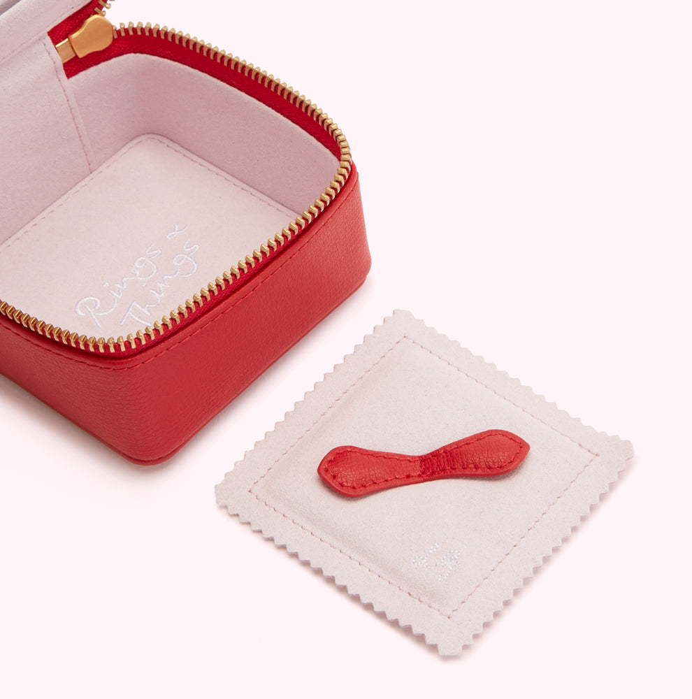 Red leather jewelry box with pale pink interior and removable red leather ring holder. The box is open, showing the interior, and the ring holder is lying beside it. The words "Rings & Things" are subtly embroidered inside the box.
