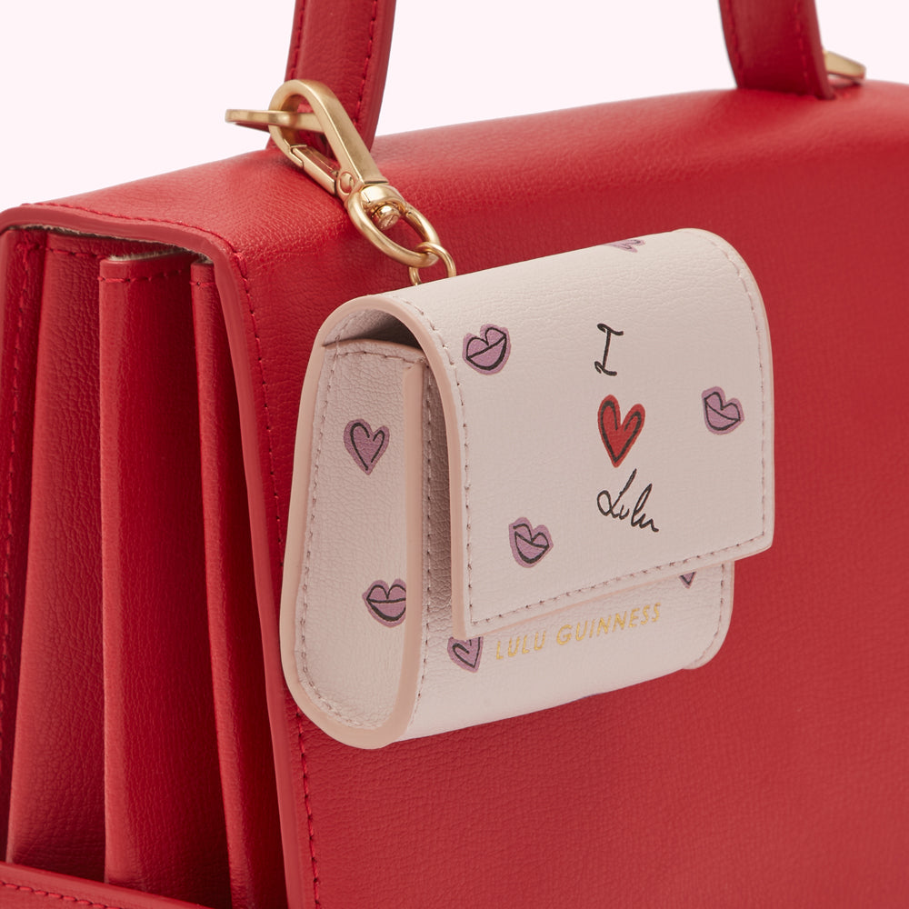 Close-up of a cream-colored Lulu Guinness air pod case with a red heart and lips design attached to a red handbag.