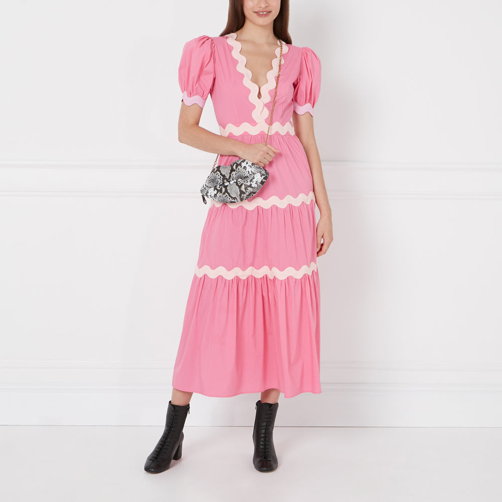 Woman in pink scalloped dress, black ankle boots, and snakeskin purse against a white background.