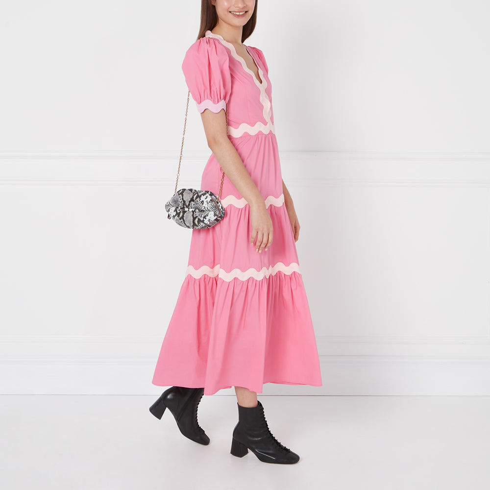 Woman in pink dress with scalloped trim and black ankle boots carrying a snakeskin purse against a white background.
