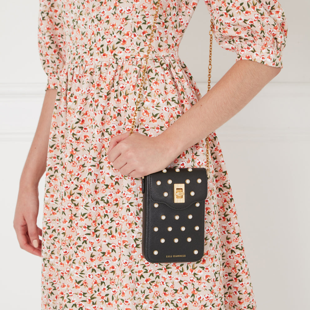 A woman in a floral dress holds a black Lulu Guinness phone case with pearl embellishments and a gold chain strap.
