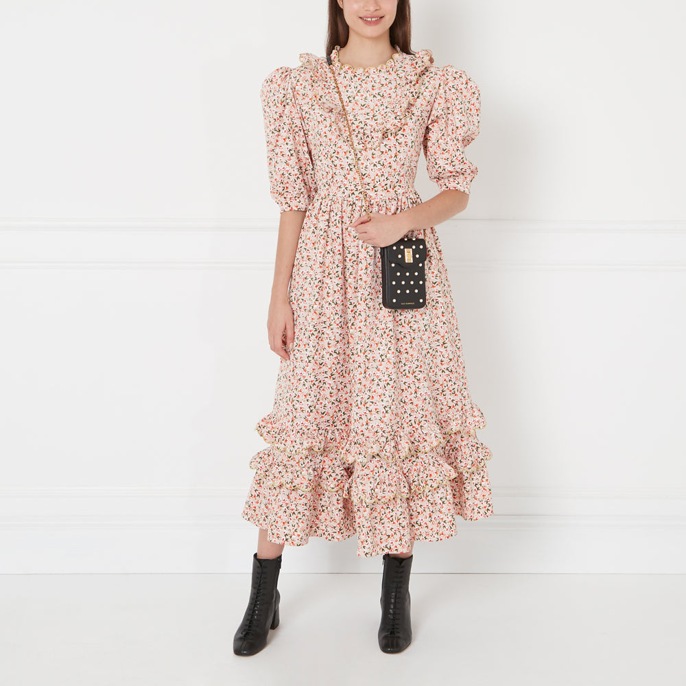 A model wearing a pink floral dress and black ankle boots carries a black studded crossbody phone bag against a white background.