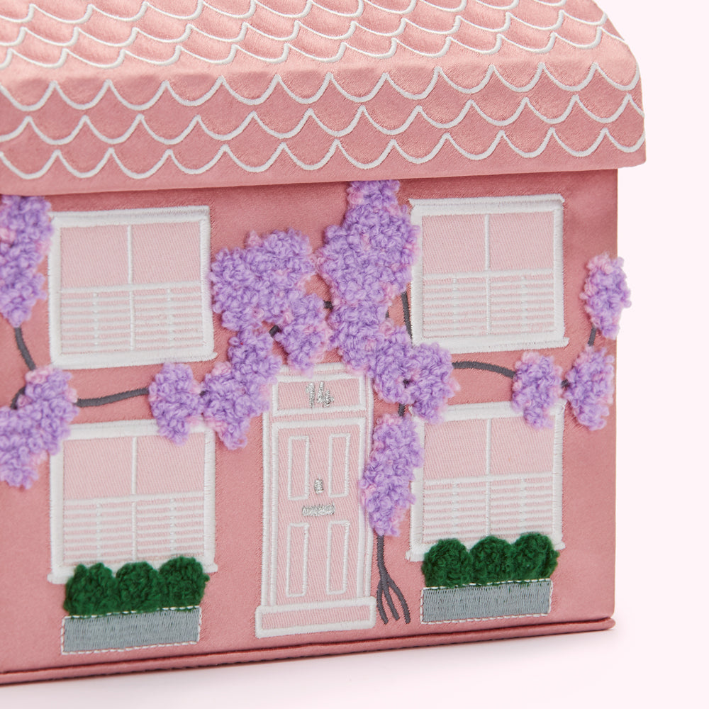 Close-up of a pink house-shaped box decorated with embroidered details, including windows, a door, and purple wisteria vines.