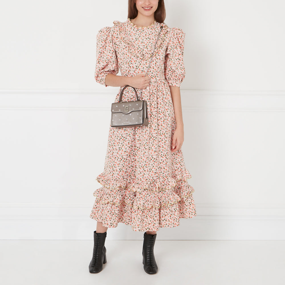 A model wearing a pink floral dress and black ankle boots carries a grey studded handbag against a white background.
