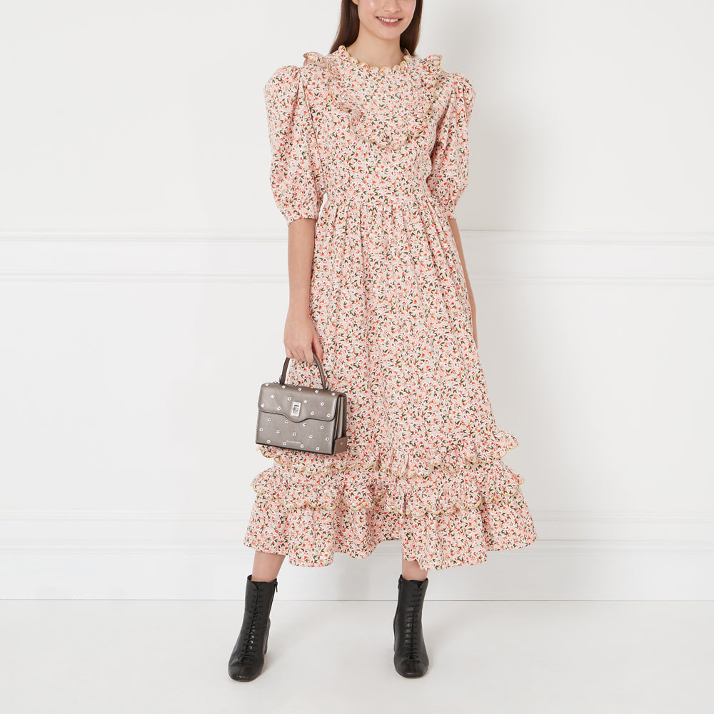 A woman in a pink floral midi dress and black ankle boots carries a gunmetal grey studded handbag against a white background.
