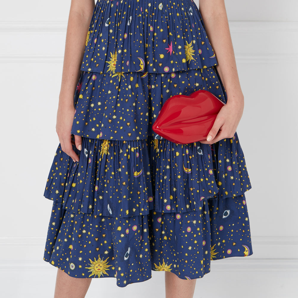 Woman in a blue tiered skirt with a celestial pattern holding a red lip-shaped clutch.
