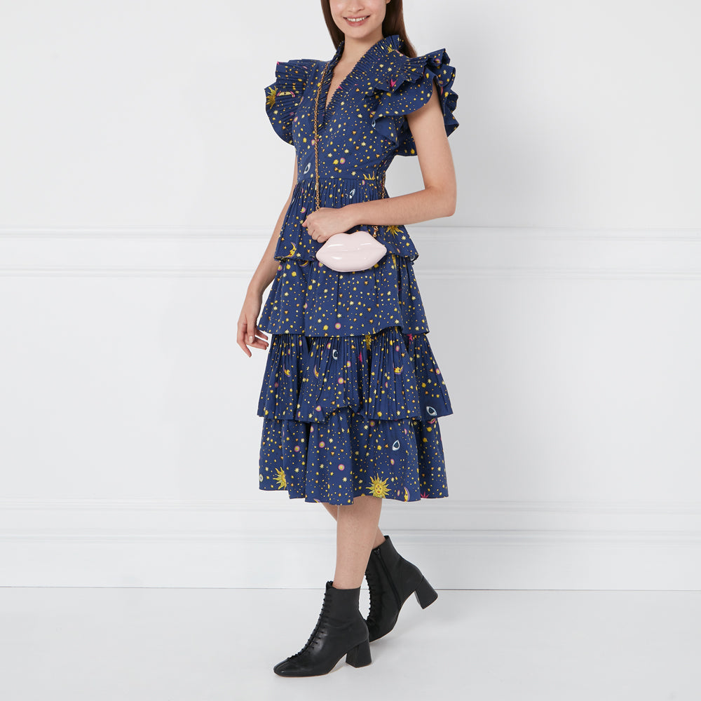Woman in a blue tiered dress with celestial print and pink lip-shaped bag, wearing black ankle boots against a white background.
