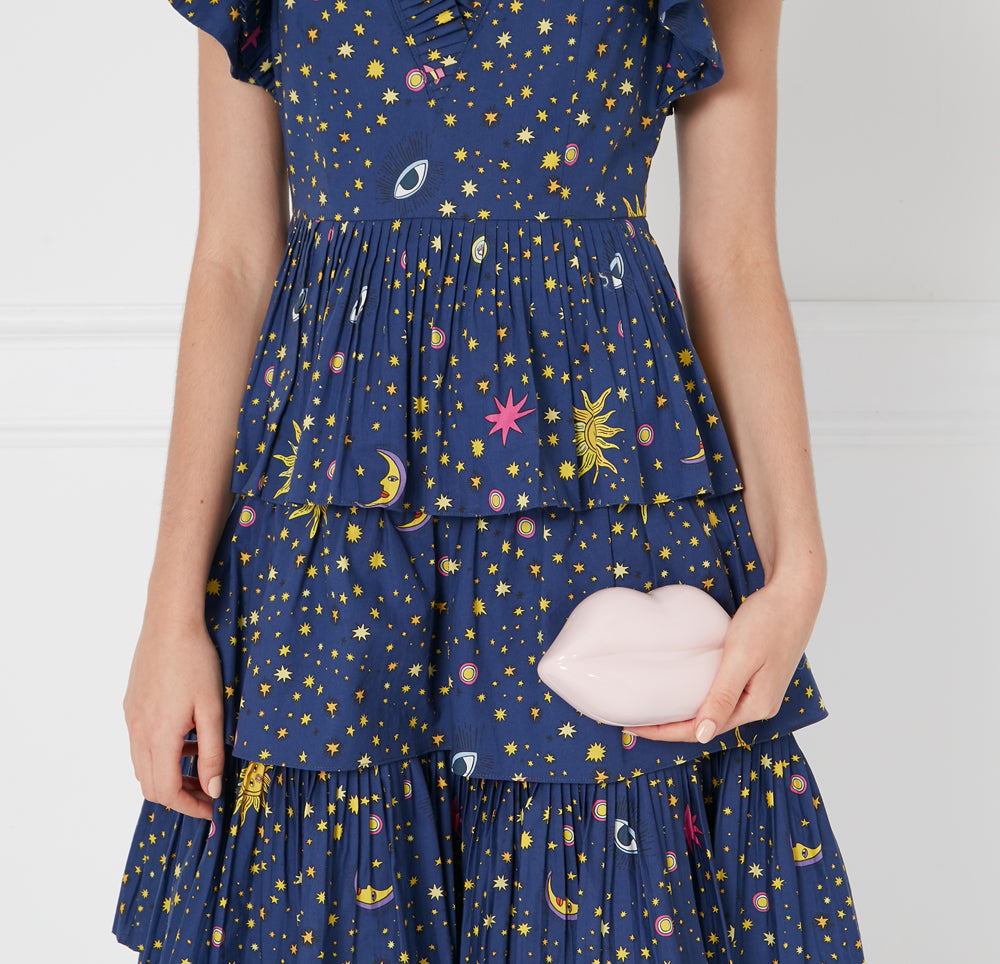 A woman in a navy blue dress with celestial print holds a pale pink lip-shaped clutch.