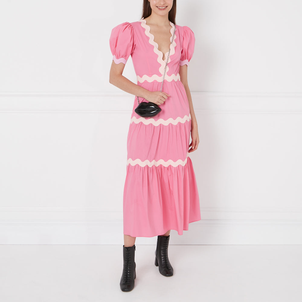 A woman in a pink dress with white scalloped trim carries a black lip-shaped purse. She's wearing black ankle boots and the background is white.
