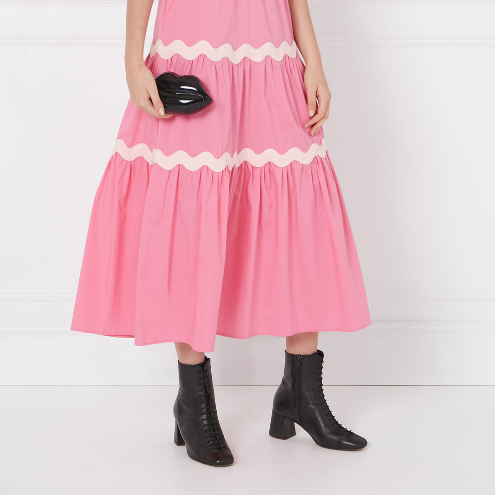 Woman in pink dress holding a black lip-shaped purse, wearing black ankle boots against a white background.
