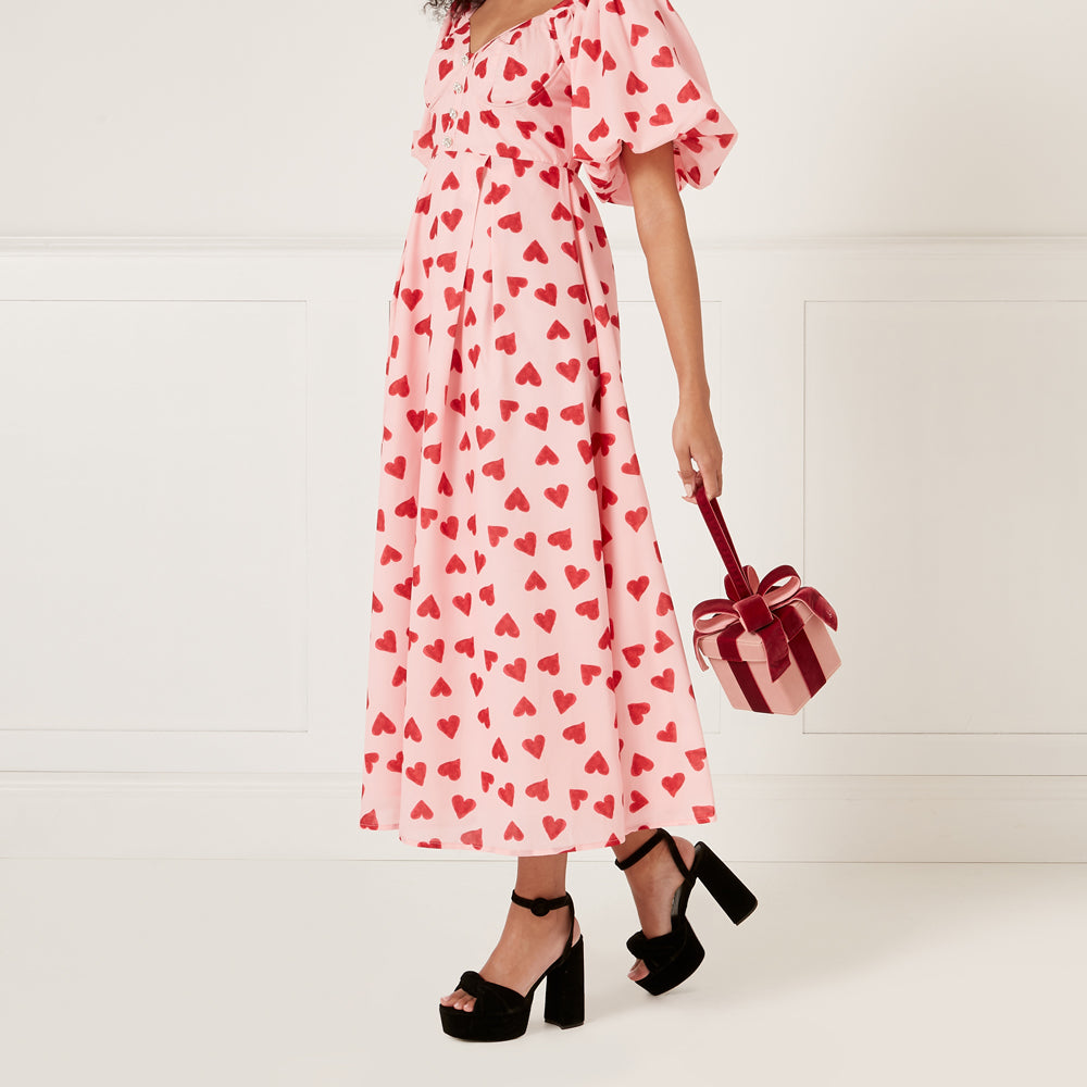 Woman in pink heart-print midi dress and black heels, carrying a small pink and burgundy gift box bag.
