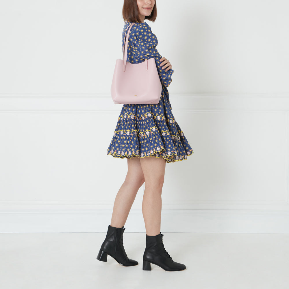 Woman in blue floral dress and black ankle boots carries a light pink tote bag.
