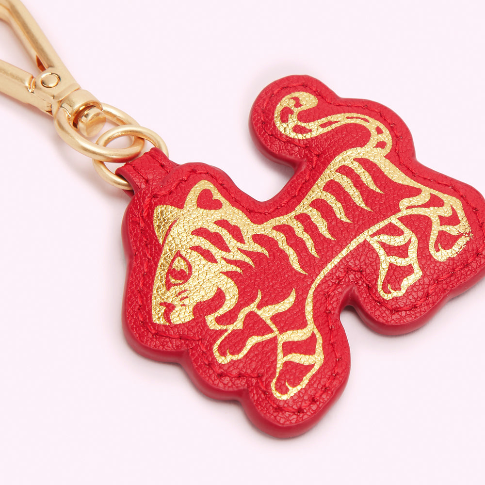 Close-up of a red leather keychain shaped like a tiger, with gold detailing on a white background.

