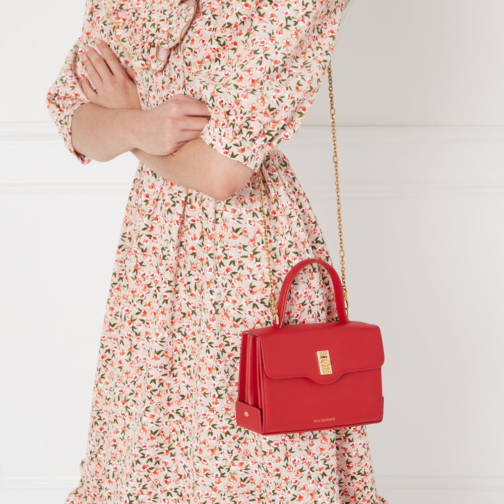 A woman in a floral dress carries a red Lulu Guinness handbag with a gold chain strap.
