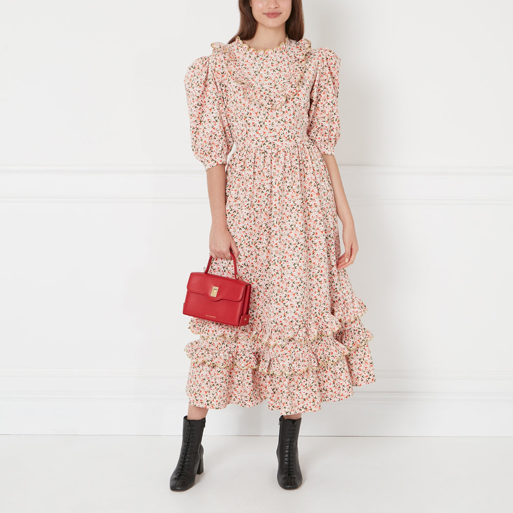 A woman in a pink floral midi dress with ruffles holds a small red handbag. She is wearing black ankle boots. The background is white.
