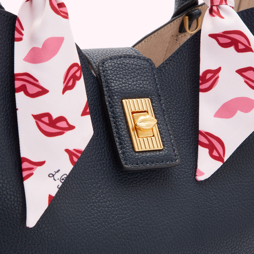 Close-up of a navy blue textured leather handbag with a gold clasp and a pink lips patterned scarf detail.
