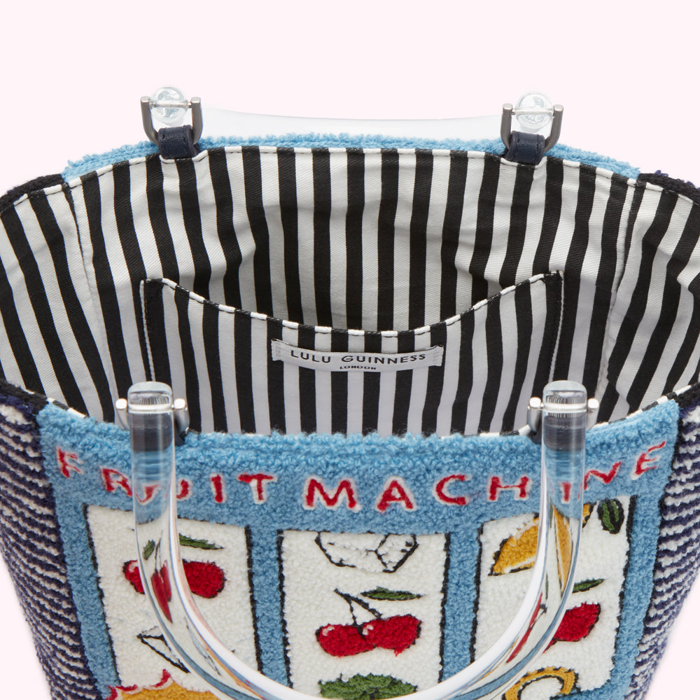 Close-up view of the inside of a Lulu Guinness London tote bag, featuring a black and white striped lining and a fruit machine design on the exterior. The bag is made of a textured fabric with embroidered cherries and other fruits on a blue background.