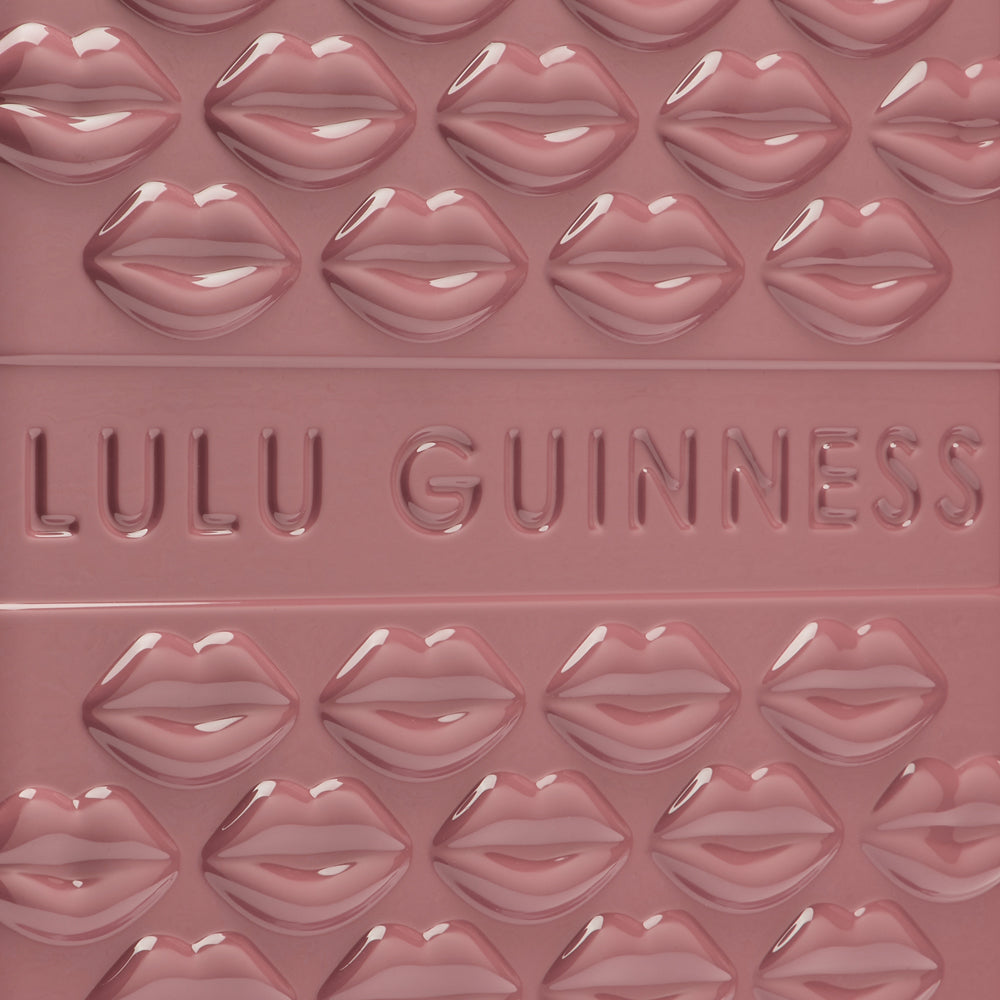 Close-up of a dusky rose Lulu Guinness product featuring a repeating pattern of embossed lips and the brand name in a recessed design.
