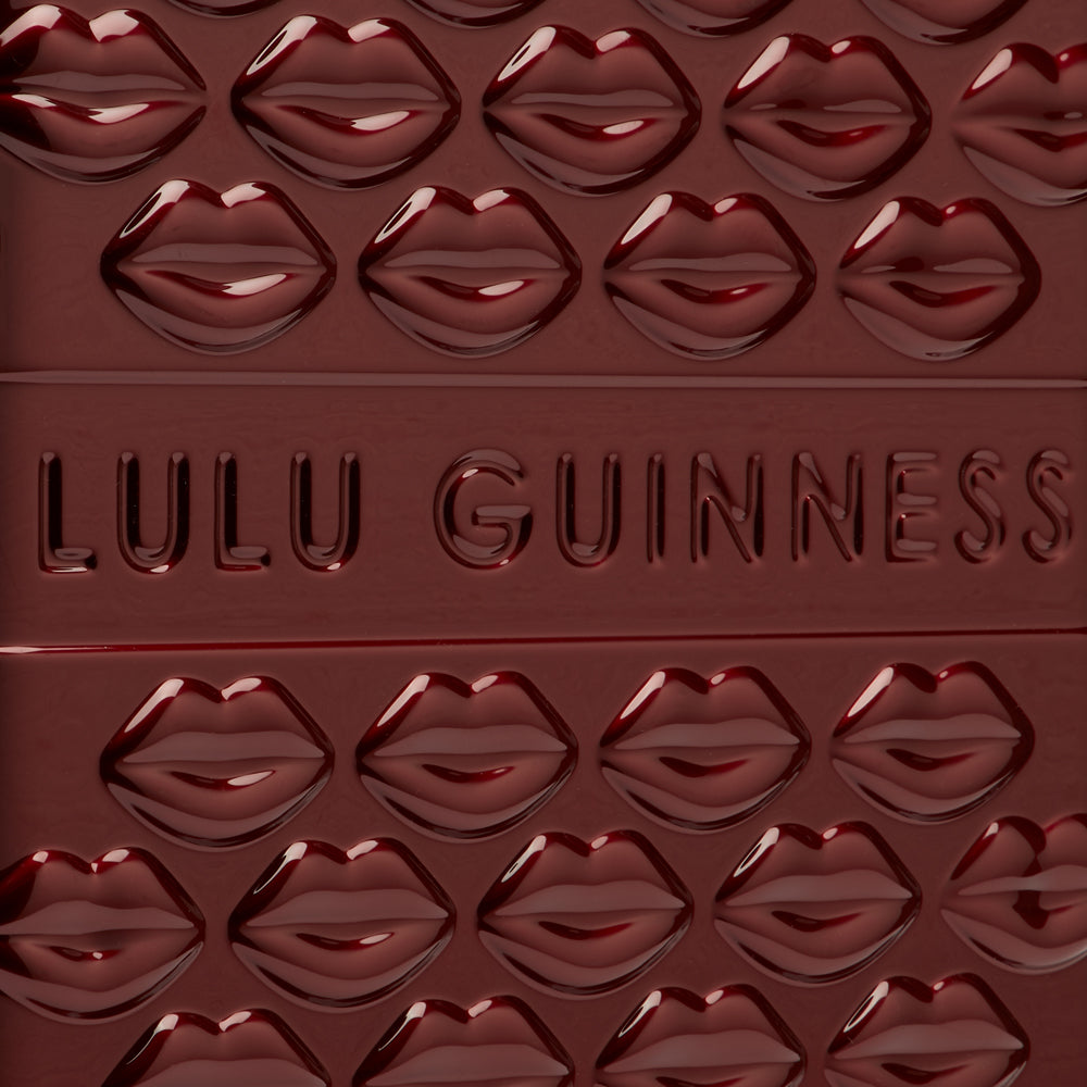 Close-up of a dark red Lulu Guinness compact with a repeating embossed lip pattern and the brand name embossed in the centre.
