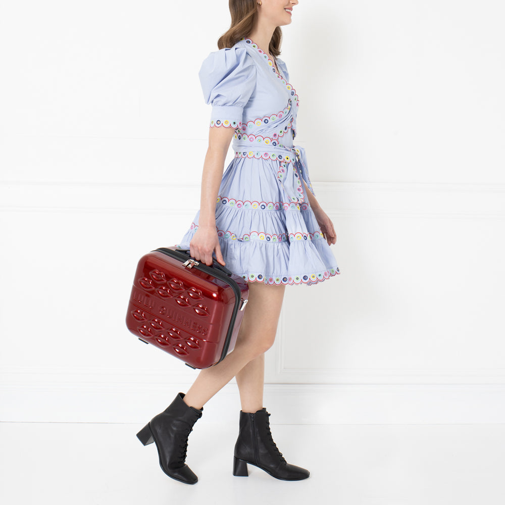 Woman in light blue dress carrying a red Lollipops suitcase against a white background.
