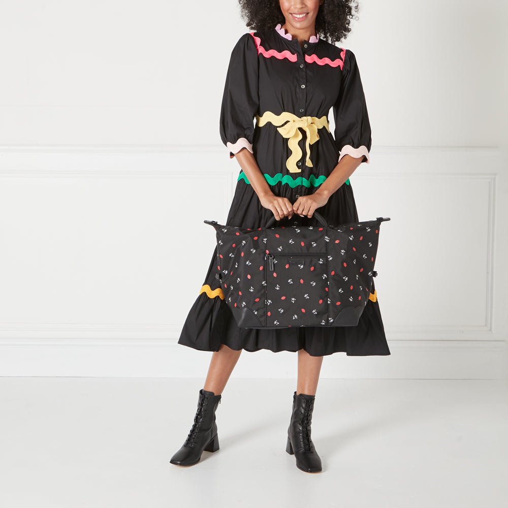 A woman in a black and colourful dress holds a black carry-on bag with a red and white lip print on a white background.
