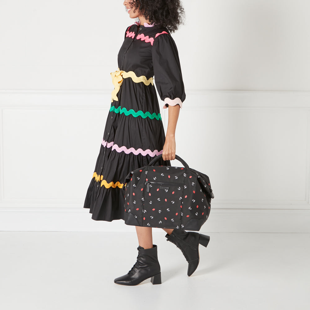 A woman in a black dress with colourful trim carries a black duffel bag with a red lips and bird pattern.