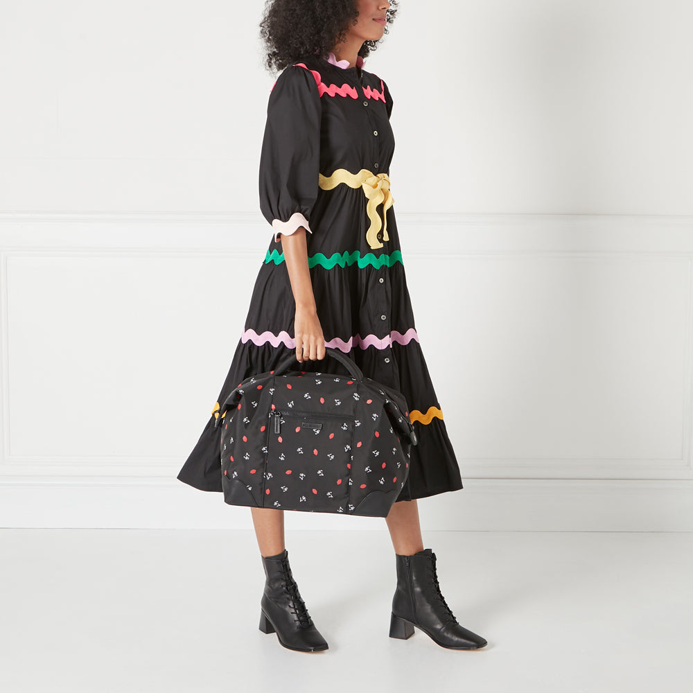 A woman in a black dress with colourful trim holds a black and red Lips print duffel bag.